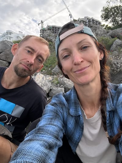 The image depicts two individuals, a woman and a man, in an outdoor setting near what appears to be a construction site and rocky terrain. The woman, positioned at the top of the frame, has fair skin, brown hair partially braided, and is wearing a light-colored headband and a blue plaid shirt over a white undershirt. She is looking directly at the camera with a slight smile. Below her, the man has light skin, short hair, and a trimmed beard. He is wearing a dark t-shirt with a blue graphic. The background includes rock formations, greenery, and buildings under construction, one with a visible crane. The perspective is angled, suggesting the photo was taken as a selfie. The presence of a modern building and a crane suggest this location may have been modernized or redeveloped in recent years. Based on these landmarks, it is not possible to pinpoint this exact location within the city of Bondarenkovo, Ukraine.