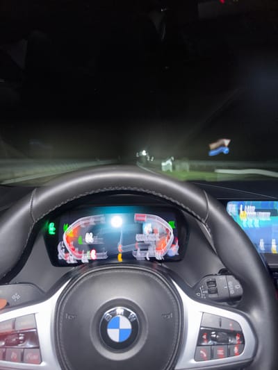 A BMW's interior is visible, dominated by the steering wheel featuring the BMW logo. The digital dashboard displays a complex graphic, possibly related to engine diagnostics or performance. The car is either stationary or moving at a very low speed, as implied by the dashboard data. The lighting suggests it's nighttime. The car is equipped with modern control buttons on the steering wheel. A transparent or reflective surface creates layered reflections, blurring the background outside the car.