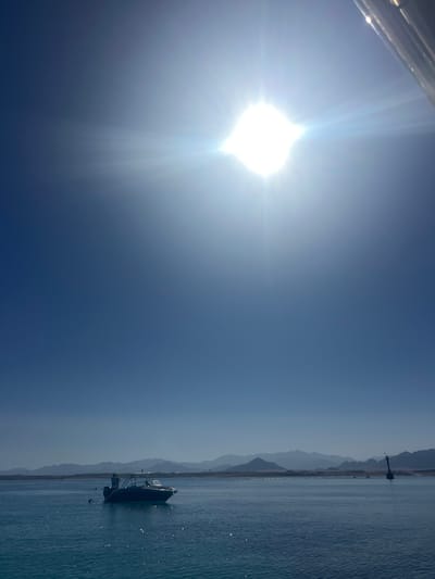 The image depicts a serene sea scene under a bright, sunny sky. The sun, a prominent bright white orb, dominates the left side of the frame, casting intense light and lens flares. The sky transitions from a bright, almost white hue near the sun to a deep blue towards the right.
In the distance, a long, thin strip of land with layered mountains is visible, suggesting a coastal or island location. The water, a deep blue, is calm with gentle ripples reflecting the sunlight, creating a sparkling effect. A small, dark boat with a person inside is anchored in the water, positioned towards the right side of the image. The boat appears to be a small motorboat. A dark, slender structure, possibly a lighthouse or a marker, stands on the distant shore.
The overall atmosphere is tranquil and suggests a clear, warm day. The time of day is likely late morning or early afternoon, given the intense sunlight and its position in the sky. No text is visible in the image. The scene evokes a sense of peace and a quiet day at sea.