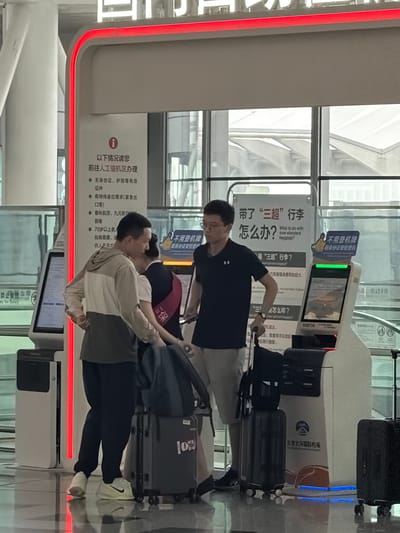 The image shows a scene inside an airport terminal, likely at Beijing Daxing International Airport in Guangyang District, China, as indicated by the text on one of the machines. Three individuals are present near self-service check-in kiosks. In the foreground, a person in a light-colored hooded jacket and dark pants is standing next to a dark gray rolling suitcase, holding the handle. To their right, another person in a dark t-shirt and light-colored shorts is partially visible, also near a suitcase. Behind them, a person wearing a dark uniform with a red sash is standing. One of the self-service kiosks features a screen with Chinese text and what appears to be an advertisement or informational display. A red illuminated strip runs along the top of the kiosk structure. The floor is reflective, indicating a polished surface. The environment appears to be a busy transit area with architectural elements suggestive of a modern airport.