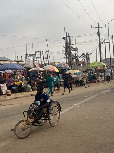 The image depicts a bustling street scene in Egan, Nigeria, under an overcast sky. In the immediate foreground, a dark-skinned male in a dark blue short-sleeved shirt and patterned trousers operates a self-propelled, three-wheeled mobility device on an asphalt road. The device features two large rear wheels and a smaller front wheel with a yellow fork. To the left, a vibrant street market occupies the roadside, characterized by multiple vendor stalls shaded by diverse umbrellas (blue, striped, green, yellow). Various goods, including produce and clothing, are displayed on tables and benches. Numerous pedestrians traverse the market area and the adjacent road. Some individuals are seen walking, while one person further back pushes a cart. The background is dominated by a complex network of utility poles, overhead electrical wires, and multiple large electrical transformers. Low-rise buildings with corrugated roofs are partially visible behind the market stalls.