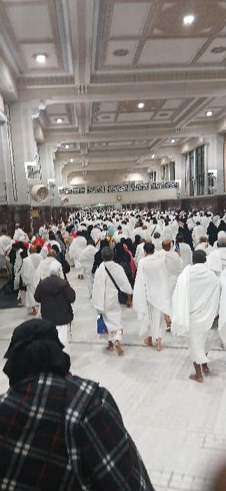 The image displays a large congregation of individuals moving through an expansive, brightly lit indoor hall located within the Grand Mosque (Masjid al-Haram) complex in Makkah Al Mukarramah, Saudi Arabia. Hundreds of people are visible, with the majority dressed in white pilgrim attire (Ihram), consisting of unstitched white cloths for men and modest white or colored garments for women. Some individuals are observed wearing darker clothing, including a person in the foreground wearing a black and white plaid jacket. The floor is light-colored, possibly marble or polished tile, and many individuals are barefoot. The hall features a high ceiling with intricate decorative patterns, recessed square panels, and integrated circular and square light fixtures. Columns or pilasters line the sides of the wide corridor. People are actively walking, generally in a forward direction, indicating movement through a designated pathway or between ritual areas. No specific interactions are discernible among individuals within the dense crowd.