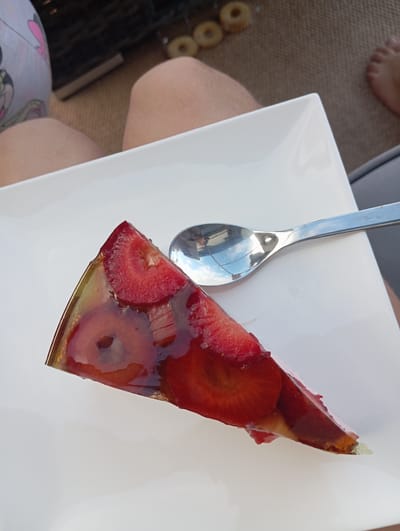 A slice of strawberry jelly cake rests on a white square plate. The cake is triangular with a translucent, amber-colored jelly covering embedded with slices of red strawberries and possibly other fruit. A silver spoon is placed to the right of the cake, reflecting the sky and what appears to be a roof. The plate is on a textured brown surface, likely a mat or rug, outdoors. In the background, a person's legs are visible, with one leg showing patterned clothing and the other bare. The scene appears to be set in Dybów Kolonia, Poland.