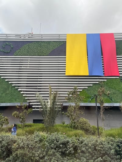 A building facade in Bogota, Capital District, Colombia, is depicted under an overcast sky. The structure features a modern design with numerous horizontal, light-colored slats and integrated decorative green wall elements, which include swirling and irregular leaf-like patterns composed of dense artificial foliage. Three large, vertical fabric banners hang prominently from the upper facade; from left to right, they are solid yellow, solid blue, and solid red, representing the colors of the Colombian national flag. The foreground and base of the building are characterized by dense, lush green landscaping, including various shrubs, bushes, and taller plants, one of which appears to be a palm. On the far left, a lone female figure with dark hair, wearing a light blue jacket or top and dark pants, is observed walking from left to right through the ground-level vegetation. A small antenna-like structure is visible on the building's rooftop.