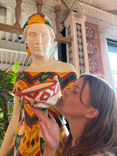 In Solnechnoye, Russia, a young woman with shoulder-length brown hair and closed eyes is leaning in to kiss a statue of a woman. The statue is pale and appears to be made of plaster, wearing a colorful dress with a geometric pattern of yellow, green, and red. The statue also sports a matching headscarf. In its hands, the statue holds a bowl decorated with red and white patterns. The background suggests an indoor environment with wooden beams and decorative plates on the wall. A large green plant is visible to the left of the statue. The lighting indicates it is daytime.