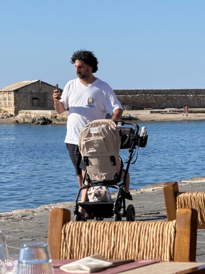 A man with curly dark hair and a beard, wearing a white t-shirt and black shorts, stands on a paved walkway next to the sea in Chania, Greece. He is holding a smartphone in his right hand and looking at it. In front of him is a beige baby stroller with a black frame. The sea is calm and blue, with a stone wall and a sandy beach visible in the background. A small stone building with a wooden door and windows is situated on the left side of the image. Two wooden chairs with woven seats are in the foreground, partially obscuring a table with glasses and napkins. The sky is clear and blue.