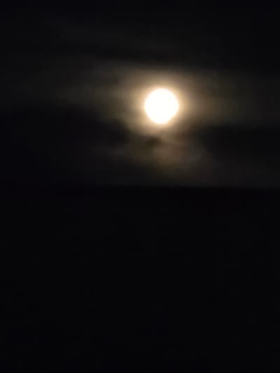 The image displays a full moon partially obscured by clouds against a dark night sky. The moon is bright and appears as a luminous orb with a hazy glow. The surrounding clouds are dark and wispy, creating a dramatic contrast with the moon's brilliance. The lower portion of the image is completely black, suggesting it may be a night landscape with no discernible features. No specific location in Буренка, Russia, can be identified from this abstract image.