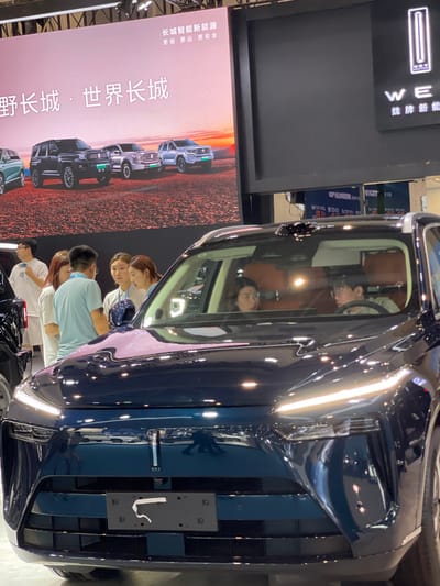 The image shows a black SUV parked in a car showroom. The car is a WEY brand vehicle. The car is surrounded by several people, including a young woman sitting in the driver's seat. Four other people are standing around the vehicle, with two looking inside and two looking away from the car. There are signs in the background advertising the WEY brand and a banner in Chinese characters. The image was taken at a car show in Hangzhou, China. The location is identified as being near the intersection of Wulin Road and Yan'an Road in Hangzhou, China, based on the provided coordinates. The image appears to have been taken indoors at a large car show.