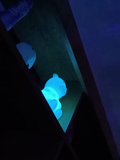 The image shows a dimly lit interior, likely a shelf unit, with a prominent blue glowing bear-shaped night light placed inside. Another smaller, similarly colored object, possibly another light, is visible on a higher shelf. A container, possibly a tube or bottle, sits on the uppermost shelf. The lighting is predominantly blue, contributing to a low-light ambiance. The overall environment suggests a residential setting, potentially a child's room. There is no information to suggest the location is in Cherkovitsa, Bulgaria. There are no people depicted in the image.