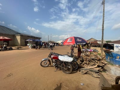 The image depicts a street scene in Bokkos, Nigeria, likely a market area. In the foreground, two motorcycles are parked on a dirt road. The one closer to the viewer is red and silver, with a white cover over its rear seat. The second motorcycle is darker and partially obscured. To the right, a pile of firewood is stacked on the ground. Behind the firewood, a stall is set up with various items, including footwear displayed on a table.
Several people are visible in the background, some standing near stalls and others near parked motorcycles or carts. A large red and blue umbrella, branded with "DANGOTE" and "YIELDS MORE," is prominent in the midground. The buildings in the background appear to be made of brick with corrugated metal roofs. A wooden utility pole with multiple wires runs vertically on the right side of the image. The sky is blue with scattered white clouds, suggesting a bright, sunny day. The overall environment is characteristic of a bustling outdoor market in a Nigerian town.