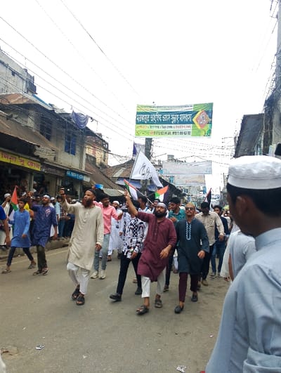 The image shows a crowd of people walking down a street in the city of Bhola, Bangladesh, at the coordinates 22.686 latitude and 90.644 longitude. The group is mostly male, dressed in traditional clothing. They are carrying flags and signs. The people are walking in a line, some are waving flags, and there are a few cars parked on the side of the street. The environment is characterized by low buildings, electrical wires, and a banner in the background. The banner appears to be advertising a local event.