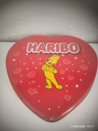 This media file shows a close-up, slightly angled shot of a red, heart-shaped tin. The tin is decorated with various small white hearts and stars. A cartoon yellow bear with a red bow on its head is prominently featured on the tin, appearing to be dancing or in motion. The tin is positioned against a white, textured background, likely a wall.
The prominent text on the tin reads "HARIBO" in large, white, capitalized letters. There is also some smaller, vertical text on the left side of the tin that is difficult to read clearly but appears to be production or ingredient information. On the right side of the image, a timestamp is visible: "17/06/2026 13:17".
The lighting suggests it is daytime, possibly afternoon, given the timestamp. The setting appears to be indoors, likely a home or a retail environment. No people are visible in the frame. The overall impression is of a decorative confectionery tin, possibly a gift item or part of a product display. The image is slightly vignette-effected, with darker edges.