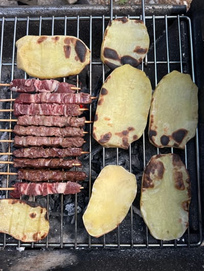 In Borno, Italy, a grill features several skewers of meat and slices of potato being cooked over charcoal. The meat skewers are arranged vertically on the left side of the grill, with each skewer containing multiple pieces of cubed meat. The potato slices are spread across the top and right side of the grill, showing signs of charring from the heat. The grill grate is made of metal, and the coals beneath are glowing, indicating an active cooking process. The overall scene suggests an outdoor cooking event or a meal preparation in a rustic setting.
