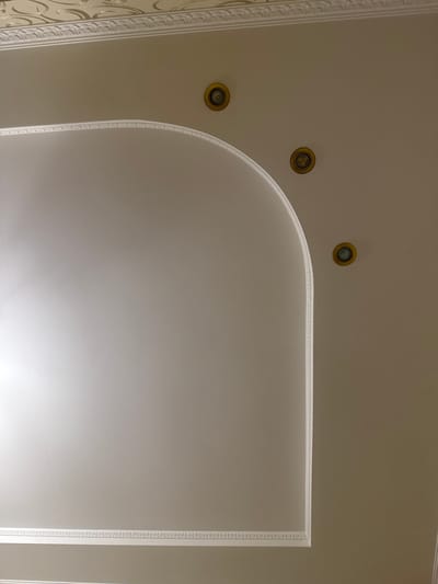The image shows a section of an interior ceiling with decorative elements. The ceiling is painted a light beige color and features a recessed area with curved edges. This recessed area is outlined with white decorative molding that has a repeating pattern. There are three circular recessed light fixtures, each with a gold-colored trim, embedded in the main ceiling area. Above the recessed section, a decorative frieze runs along the top edge of the image, also in white. The lighting in the room appears to be coming from the recessed lights and possibly from an unseen source, casting soft shadows and highlighting the textures of the ceiling and molding. The overall impression is one of a well-decorated interior space, possibly a living room or dining area. The image does not contain any people or signs of human activity beyond the installed fixtures and decorative features. The location in Гожули, Ukraine, is not discernible from the visual elements of the image itself.