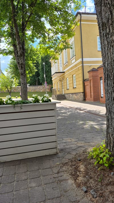 The image captures a sunlit outdoor scene on a cobblestone walkway in Zarasai, Lithuania. In the foreground, a white wooden planter box is filled with small white flowers and green foliage. To the right, a large tree trunk is visible. Behind the planter, a yellow building with white trim and windows stands prominently, featuring a balcony with white wrought-iron railings. Red brick elements, including a pillar and wall, are integrated into the building's facade. A set of stairs leads to an entrance on the building's ground floor. Beyond the building, lush green trees and a stone wall line a grassy area. The sky above is a clear blue with scattered clouds. A person can be seen walking in the distance on the left side of the frame. A sign with text and an arrow is affixed to the red brick structure. The overall environment suggests a well-maintained urban or public space.