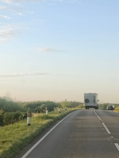 The image depicts a serene, open road scene in East Suffolk, United Kingdom, captured from a moving vehicle. The asphalt road, marked with a solid white line on the left edge and broken white lines in the center, curves gently to the right as it extends into the distance.
Ahead, a large, grey motorhome or RV is prominent, viewed from its rear. It features a spare tire mounted on the back and a ladder on its left side. Further down the road and slightly to the right of the motorhome, a dark-colored car, possibly a hatchback, is also traveling in the same direction.
The left side of the road is bordered by a grassy verge interspersed with white and orange-topped reflective road markers. Beyond the verge, dense green foliage, including bushes and trees, lines the horizon. Faint outlines of power lines or utility poles are visible in the distant haze above the trees.
The sky is a clear, light blue at the top, gradually transitioning to a pale, warm yellow-orange closer to the horizon, suggesting a late afternoon or early evening setting. A few wispy clouds are scattered across the sky. The overall impression is one of peaceful travel through a rural or semi-rural landscape. There is no visible text in the image.