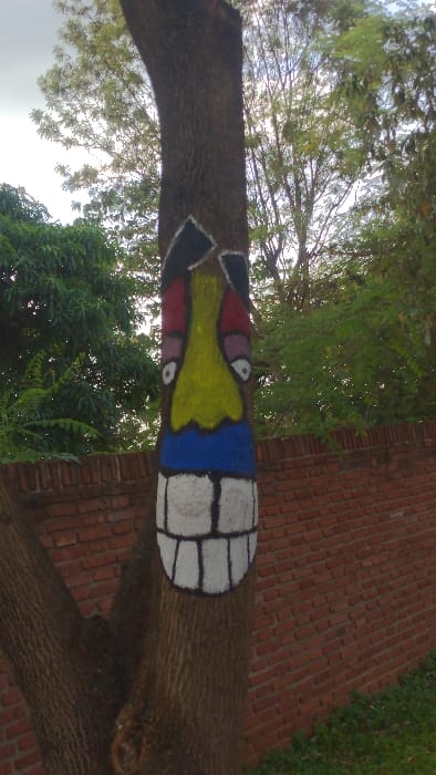 A tree trunk, oriented horizontally, features painted art. On the left, a black and yellow design surrounds an eye. The trunk transitions into shades of red and yellow, also encompassing another visible eye. Blue paint follows, ending with a pattern of large white squares outlined in black. Beyond the painted section, the natural wood bark is visible. Lush green trees and foliage surround the tree trunk, with a brick wall appearing in the background.