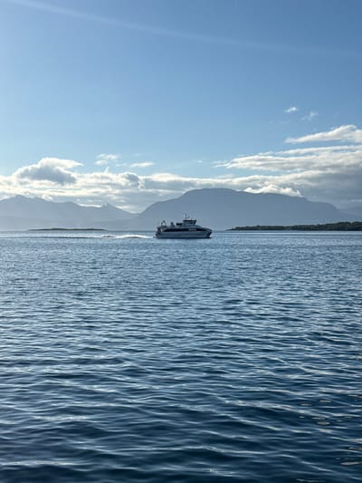 A ferryboat is pictured in the distance, cruising on the water under a clear blue sky with scattered clouds. Majestic mountains form the backdrop, their peaks shrouded in a soft haze. The water is calm, with gentle ripples reflecting the sunlight. The scene appears to be a tranquil moment captured in the natural beauty of Harstad, Norway.