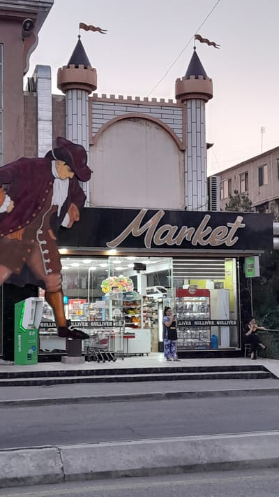 The image shows a market in Bukhara, Uzbekistan. A large, stylized cutout of a man in historical attire is positioned to the left of the market's entrance. The market sign, which reads "Market" in large, cursive lettering, is prominently displayed above the entrance. The market appears to be a convenience store or small grocery store, with shelves stocked with goods visible through the glass storefront. Two women are standing in front of the market. One woman, on the right, is seated and looking away from the camera. The other woman, in the center, is standing and looking at her phone. The street in front of the market is visible, with a sidewalk separating it from the building. The architecture of the building features decorative elements resembling a castle, including turrets and crenellations. The sky above is a pale, overcast gray, suggesting it might be early morning or late evening. The overall scene captures a typical street view in a commercial area of Bukhara.