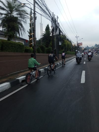 The image depicts a street scene in Dawathang_Dorjibi_ Kashingtsawa, Bhutan, under an overcast sky. The asphalt road surface is wet, suggesting recent precipitation.

In the foreground, three individuals are riding bicycles on the left side of the road, adjacent to a black and white striped curb. The leftmost cyclist wears a green long-sleeved shirt and dark shorts, riding a bicycle with red wheel rims. The second cyclist, positioned slightly to their right and behind, wears a black t-shirt with a graphic design and dark shorts, riding a black bicycle. The third cyclist, further to the right and slightly behind, also wears a dark t-shirt with a graphic and dark shorts, riding a black bicycle.

Further down the road, to the right of the cyclists, two motorcyclists are visible. The first motorcyclist wears a grey hooded garment and dark trousers. The second, further back, wears a light-colored jacket. Additional motorcycles and vehicles are discernible in the distance.

On the left side of the frame, a raised embankment features a brown retaining wall topped with trimmed green bushes. Behind the foliage, a building with a white facade and dark roof is partially visible. Several tall palm trees and evergreen trees populate this area. A prominent utility pole with a dense network of overhead wires stands near the curb, with additional poles and wires extending into the background on the right. Decorative lights are visible on a vertical element behind the primary utility pole on the left.

In the mid-ground on the right, a street light fixture is visible, along with a commercial sign displaying the name "ALFAMART." The background shows hilly terrain with scattered trees and structures, receding under the uniform grey sky.