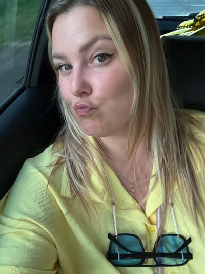 The media file shows a close-up, slightly tilted portrait of a young woman. She has blonde hair with lighter streaks and brown eyes. Her expression is a pout, with her lips pursed. She is wearing a bright yellow, collared shirt. A delicate gold necklace is visible around her neck. Black-rimmed sunglasses with blue lenses are hanging on a beaded white string, resting on her shirt. The background appears to be a car interior, with a dark seat on the left and a window with a blurry green landscape outside on the right. The lighting suggests it could be daytime. There is no visible text in the image. The overall impression is a casual, self-taken photograph, possibly in a vehicle during daylight hours, with no specific event or activity clearly indicated beyond the posing for the photo.