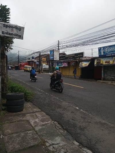 This image depicts a street scene in Neerveli, Sri Lanka, under an overcast sky. The view is from a low angle on the right side of an asphalt road, looking forward and slightly uphill.
In the immediate foreground, on the left, a stack of three used vehicle tires rests on a concrete sidewalk slab next to a patch of green vegetation. The sidewalk exhibits cracks and discoloration.
The asphalt road, dark gray with some visible wear, features a faded yellow double line marking in the center-right. Two individuals are operating motorcycles on the road. The first motorcyclist, positioned further back and slightly to the left, wears a blue jacket and blue helmet, riding a smaller, dark-colored motorcycle. The second motorcyclist, closer to the viewer and in the center of the road, wears a brown jacket and a light blue helmet, carrying a dark backpack, and is riding a dark scooter.
Along the right side of the road, a row of single-story commercial buildings or shops is visible. Many storefronts appear closed, some with yellow or dark roll-down security gates. Above these buildings, numerous utility poles are visible, densely strung with a multitude of overhead electrical or communication wires. Several large commercial signs are mounted on the buildings, including one with "Bollywood" written in white lettering and other text that appears to be in Arabic or Jawi script. Another prominent sign on the far right advertises "Crystal" beverages and "TOKO ALA".
Further down the street on the left, a yellow van is parked or moving slowly. A pedestrian in dark clothing is visible walking on the sidewalk near the closed storefronts on the right.
On the far left, a white rectangular sign with dark lettering, reading "Spooring & Balancing", is mounted on a pole or tree trunk. Lush green foliage from a tree is partially visible around this sign. In the distant background, hazy hills or mountains are discernible beneath the cloudy sky.