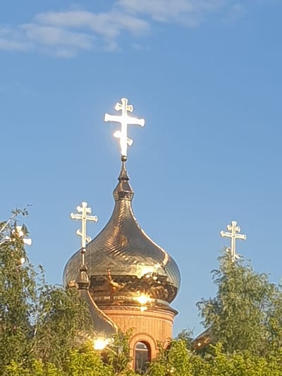 This is a close-up, eye-level, vertical, daytime shot of an Orthodox church dome topped with a golden, multi-tiered cross. The dome is made of reflective gold material and is surrounded by lush green trees. A bright blue sky with scattered white clouds serves as the background. The church appears to be the dome of the Church of the Intercession of the Most Holy Mother of God, located in Obukhiv, Ukraine.