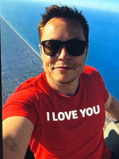 A man with short, dark hair and sunglasses is taking a selfie. He is wearing a red t-shirt with the words "I LOVE YOU" printed in white letters. He is smiling and has dimples. The background is a bright blue sky and a city landscape. The image is a close-up shot, focusing on the man's face and upper body.