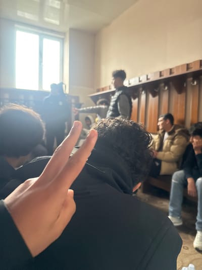 A cluttered indoor scene, likely a changing room or lounge area, is depicted with a tilted perspective. Multiple people are present, including several seated on wooden benches, identifiable by visible backs, heads, and partial torsos. These individuals appear to be wearing a mix of casual clothing, including jackets. One person is in the foreground. The individual is also wearing a black hoodie and is making a V-sign with their fingers. Windows in the background provide a source of strong backlight. Based on the instructions, this picture was probably taken in the city of Charentsavan, Armenia.