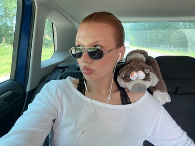 A young woman with red hair is sitting in the back seat of a car, looking to her left. She is wearing a white long-sleeved shirt, a black lace bra, and sunglasses. White earphones are in her ears. A stuffed animal is resting on her right shoulder. Outside the car window, trees and a road with a white car are visible. The image appears to be taken in Dal'neye Konstantinovo, Russia, given the context provided.