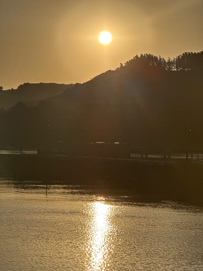The image depicts a serene waterscape with a dramatic sunset over Zumaia, Spain. The sun, a bright orb, hangs in the hazy sky, casting a warm, golden glow over the scene. Silhouetted against the radiant sky are rolling hills covered in a dense forest of pine trees. In the middle ground, a bridge spans across a body of water, its structure partially obscured by the low light. A few indistinct figures are visible on or near the bridge, adding a sense of scale to the vastness of the landscape. The foreground is dominated by calm water, its surface shimmering with the reflection of the setting sun, creating a dazzling display of light and color. The overall atmosphere is one of tranquility and natural beauty, capturing a moment of quietude as day transitions into night.