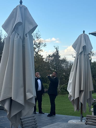 Two men dressed in suits are standing on a stone patio, possibly outside. One man is holding a wine glass and appears to be drinking from it. Behind them are trees and a grassy area. Above and below them are closed, beige umbrellas. The location is unknown beyond being in Stellenbosch Nu, South Africa.