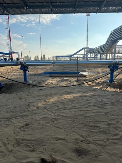 The image depicts an industrial construction site under a clear blue sky with scattered clouds. In the foreground, a sandy ground is covered with numerous footprints, suggesting recent activity. A large, silver-colored pipe runs horizontally across the mid-ground, supported by blue metal structures. To the left, a red and white striped tower is visible, and further back, more industrial infrastructure, including elevated pipelines and other towers, can be seen. The environment appears to be an open area with a few distant trees on the horizon. The overall scene conveys a sense of ongoing industrial development in a relatively undeveloped area, consistent with the location in Chikshino, Russia, which is known for its oil and gas infrastructure development.