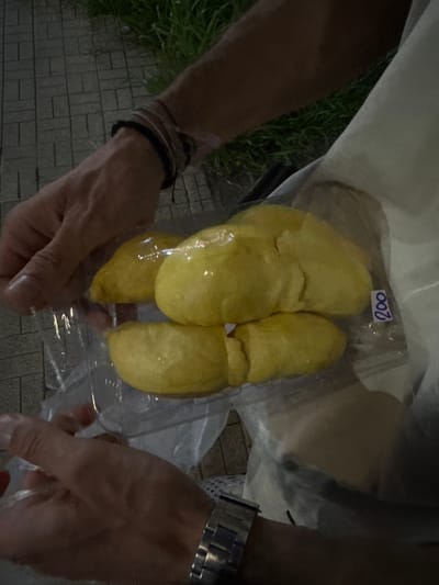 In an outdoor setting with a cobblestone walkway and grass in the background, a person is holding a clear plastic bag containing several pieces of yellow durian. The bag is sealed and has a blue sticker with the number "200" written on it. The person's hands are visible, with one hand holding the bag from the top and the other supporting it from the bottom. The person is wearing bracelets on their left wrist and a watch on their right wrist. The lighting suggests it is evening or night. The location is identified as Surat Thani City Municipality, Thailand.