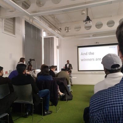 The image depicts an indoor presentation or event held in a modern lecture or multi-purpose room at Coventry University in Coventry, United Kingdom.
At the front of the room, a bald, dark-skinned male, dressed in a dark suit jacket and a white shirt, stands facing the audience. He is positioned in front of a large white projection screen which displays the text "And the winners are." in grey font, with the "Coventry University" logo visible in the upper right corner of the screen.
An audience is seated on grey chairs with metal frames, arranged in rows on a green carpeted floor, facing the speaker and screen. The immediate foreground shows the back of an individual wearing a blue and white checkered shirt and a grey baseball cap with the number "1994" embroidered on the back. To their left, another attendee is visible from the back, wearing a dark top and blue jeans. Further left, near a wall with large windows covered by horizontal blinds, two individuals are present; one is standing behind a black video camera mounted on a tripod, while another is seated nearby.
The room features white walls and a ceiling with exposed white structural beams containing circular cutouts, from which various light fixtures and a ceiling-mounted projector extend. A white table is located in front of the speaker.