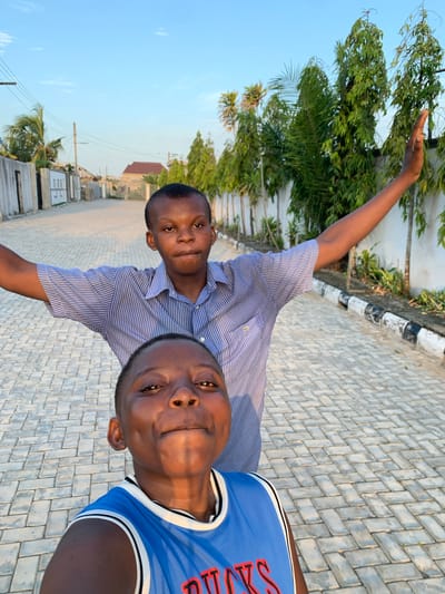Two young males are featured in a bright outdoor selfie-style photo, likely taken on a paved street in Ibasa, Nigeria. The person in the foreground, positioned on the right, wears a blue sports jersey with white and black striped trim around the neck and sleeves. Partially visible red text on his jersey reads "RS". He looks directly at the camera with a neutral to slightly smiling expression.

Behind and slightly to the left, the second person wears a blue and white striped short-sleeved button-up shirt. He has his right arm raised and slightly bent, with his hand visible against the clear sky and foliage, also looking towards the camera with a calm expression.

The setting is an outdoor area with a grey paved surface, likely a street or pathway, made of interlocking blocks. A black and white painted curb runs along the left side of the paving. The background features abundant green trees and foliage, indicating a natural or semi-urban environment. Beyond the trees, the horizon reveals houses, including one with a reddish-brown roof, and utility poles with power lines against a clear, light blue sky. The warm, soft lighting suggests either late afternoon or early morning. The overall mood is casual and relaxed.