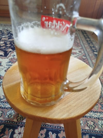 A close-up, slightly elevated shot shows a mug of amber-colored beer filled to the brim, with a thick layer of white foam. The mug is resting on a round, light-brown wooden stool, which is placed on a patterned carpet with navy blue, pink, and white floral motifs. The background is blurred, showing what appears to be a wooden surface and a red label on the mug with white text. The scene appears to be indoors, possibly in a domestic setting, within the city of MZ Petrov Gaj, Bosnia and Herzegovina. There are no people or active interactions depicted in the image.