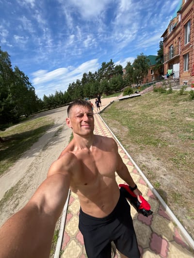 In Lebyazhye, Russia, a shirtless man takes a selfie outdoors on a sunny day. He is wearing black shorts and holding a red and black item in his right hand. The man is standing on a patterned tiled pathway. Behind him, a dirt path runs parallel, bordered by grass and trees. In the background, there are several brick buildings with green roofs and a clear blue sky with scattered white clouds. Other people are visible in the distance, walking along the paths and near the buildings.