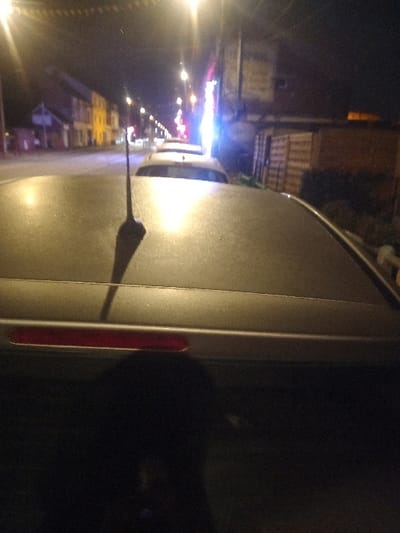 A nighttime view of a vehicle's side, possibly a van, dominates the frame. The street stretches out horizontally in the background. Lights illuminate nearby buildings and traffic. The vehicle is silvery-gray, with a prominent side mirror casting a sharp shadow onto the panel. A red reflector is visible near the van's rear. The image appears to be taken from a slightly elevated position, accentuating the urban environment.