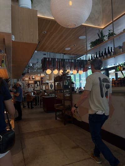 The image shows the interior of a restaurant or cafe, likely taken in Austin, United States of America, as inferred from the architectural style and restaurant ambiance typically found in the city. A man in a white t-shirt and jeans is captured, possibly holding a package or object, situated towards the middle-right of the frame. There appear to be other patrons within the establishment, with some seated at tables. The background showcases artwork, including framed pieces, and a distinctive lighting fixture. The presence of bottled beverages along the upper part of the frame suggests an emphasis on culinary offerings and drinks. The setting seems to be a casual yet upscale dining place with a blend of modern design and local art. The photo appears to have been rotated.