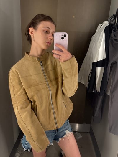 A young woman is taking a mirror selfie in what appears to be a fitting room or a small, private space. She is wearing a tan suede jacket with a zippered front and a collared neckline, paired with distressed denim shorts. Her hair is pulled back, and she is wearing a small earring in her left ear and rings on her fingers. She is holding a light purple phone with a speckled case to take the picture. Behind her, to the right, are several garments hanging on a rack, including a white and black jacket and a gray garment. The walls of the space are a muted brown color. There is a clear plastic cup on the floor to the left of the woman. No specific location within Aschaffenburg, Germany can be identified from the image.