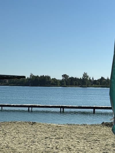 The image depicts a serene lakeside scene. In the foreground, a sandy beach with visible ripples and texture stretches out, suggesting it is dry. A wooden pier extends into the clear blue water, supported by pilings. The water is calm, with subtle wave patterns. Across the lake, a dense line of green trees forms a natural barrier, with some buildings partially visible behind them. The sky is a clear, bright blue. The overall impression is one of a peaceful, sunny day by the water. The location is identified as the city of Verbizh, Ukraine.