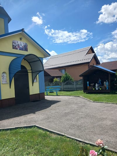 The image depicts a small, bright yellow and blue building with a dark door and an arched blue awning in Doslidnytske, Ukraine. Above the door, a mural depicts a religious scene. To the left of the entrance, a small niche contains an icon. A blue bench sits on the paved area in front of the building. Further back, a wooden structure with a blue roof houses a nativity scene with figures of Mary, Joseph, baby Jesus, sheep, and angels. Behind the nativity scene is a grey concrete fence and a brown-roofed house. The sky above is blue with scattered white clouds. The ground in the foreground is covered in green grass and a paved pathway. Two pink roses are visible in the lower right corner.