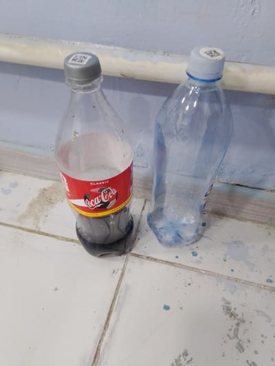 Two plastic bottles are placed on a tiled floor next to a wall in Nurafshan (Toytepa), Uzbekistan. The bottle on the left is a Coca-Cola bottle, half-filled with a dark liquid, and has a QR code on its grey cap. The bottle on the right is a clear plastic bottle, empty except for a small amount of water at the bottom, and has a white cap with a blue ring and a QR code. A light-colored pipe runs horizontally along the wall above the bottles. The tiled floor appears to have some blue and brown stains.