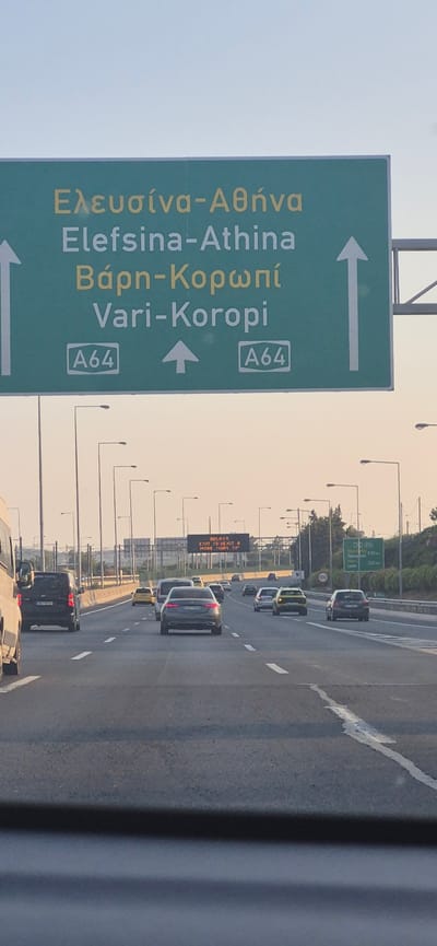The image shows a highway scene in Paiania Municipal Unit, Greece, featuring a large green overhead road sign. The sign displays directional information in both Greek and English, indicating routes to Elefsina-Athina, and Vari-Koropi, with associated highway numbers "A64". Arrows on the sign point straight ahead and to the right, suggesting different directions for the listed destinations. The highway itself has multiple lanes with cars driving in them. In the distance, another digital sign is visible, displaying a message in red text about "DELAYS" and "EXIT 15 < EXIT 8 MORE THAN 30'". The sky is clear and appears to be late afternoon or early evening. Streetlights are present along the side of the road.