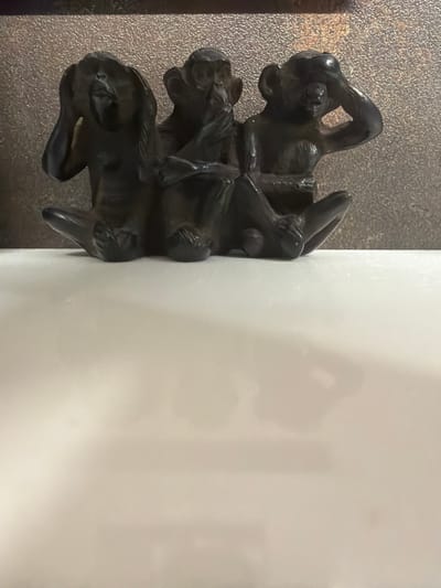 The image shows a decorative element consisting of three stylized animal figures stacked vertically, likely made of dark metal. The figures appear to be stylized depictions of cats or similar creatures. The element is attached to the edge of a white object, possibly a piece of furniture or architectural feature. The background suggests a surface with a textured or granular appearance. The photograph was taken in Arinj, Armenia.
