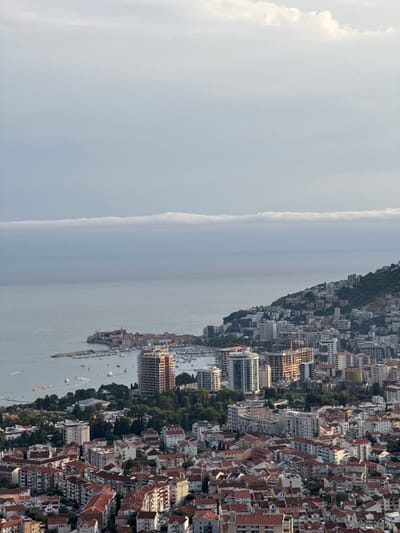 This image is an aerial view of the coastal city of Košljun, Montenegro. The scene captures a dense urban landscape with a variety of buildings, ranging from older structures with red-tiled roofs to modern high-rise apartments and hotels. The city is situated on a hillside that slopes down towards the Adriatic Sea. Several boats are visible in the harbor, and the sea stretches out to the horizon, where a distinct, thin layer of stratocumulus clouds is present. The sky above is a mixture of lighter and darker shades of gray and white, suggesting overcast conditions or the approach of evening. The overall impression is of a bustling Mediterranean city with a backdrop of natural beauty.