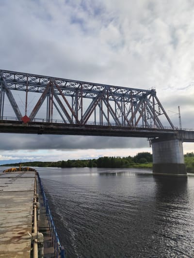 A photograph depicts a steel truss bridge spanning a river. The bridge structure shows some rust. The sky above is overcast. In the foreground, the flat deck of a vessel is visible, with blue railing extending along the side. The river surface shows ripples and waves. Trees and vegetation line the distant shoreline. The scene is located in Tarkanovo, Russia.