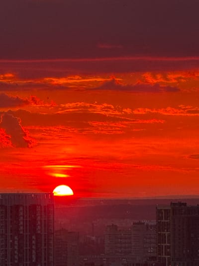 The image captures a vibrant sunset over the city of Savenki, Russia. The sky is ablaze with intense shades of orange and red, with scattered clouds illuminated by the setting sun. The sun itself, a brilliant orb, is partially obscured by the horizon, casting a warm glow. In the foreground, the silhouettes of modern buildings rise against the dramatic sky, indicating an urban setting. The overall scene conveys the dramatic beauty of a sunset in a city environment.
