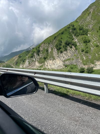 The image captures a view from a vehicle traveling along a mountainous road in Almasiani, Georgia. The foreground shows a gravel road and the side of a car. A silver guardrail runs alongside the road, separating it from a steep, grassy mountainside dotted with rocks and trees. The mountain is covered in lush green vegetation with distinct rocky outcrops. In the distance, more mountains are visible, partially obscured by clouds. The sky is partly cloudy. A side-view mirror of the car reflects a person holding a phone, presumably taking the picture. The scene is natural and appears to be a scenic route.